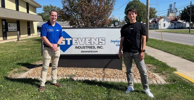 Curtis Garrard and Volodymyr Hresko standing either side of the Stevens Industries, Inc. sign outside the company's Illinois plant